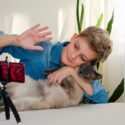 En dreng vinker til en smartphone, der står på et stativ, mens han holder en kat i armene; det ser ud til, at han foretager et videosamtale eller optager en video i et lyst rum med planter i baggrunden.