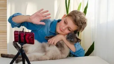 En dreng vinker til en smartphone, der står på et stativ, mens han holder en kat i armene; det ser ud til, at han foretager et videosamtale eller optager en video i et lyst rum med planter i baggrunden.