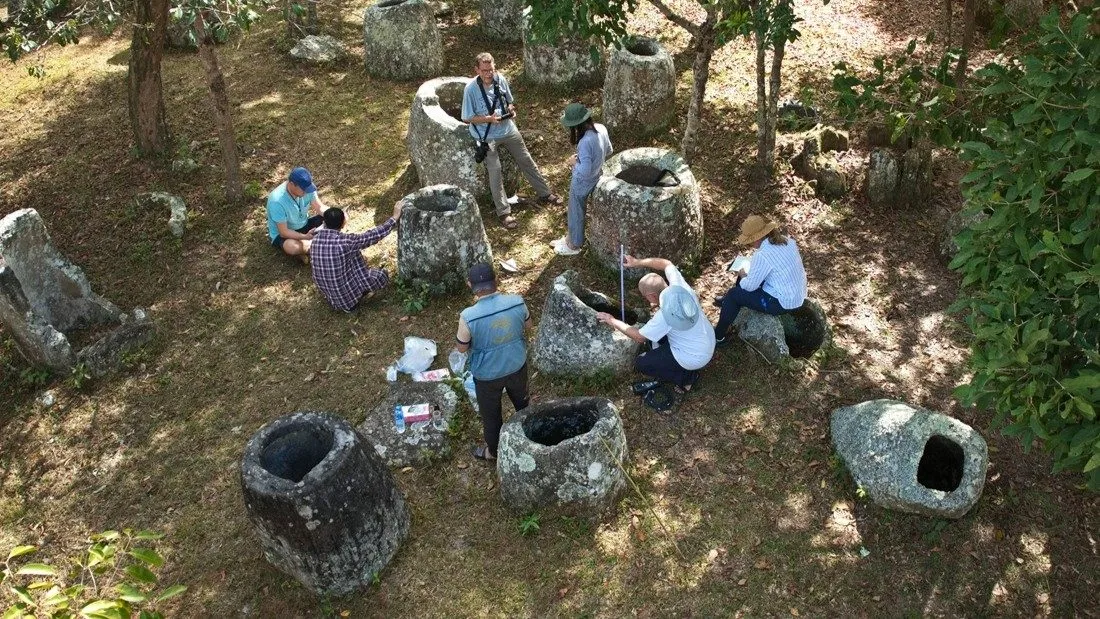 En gruppe mennesker undersøger og sidder omkring store, gamle stenkrukker på en græsklædt udendørs plads omgivet af træer. Der ligger arkæologiske redskaber og notesbøger på jorden.