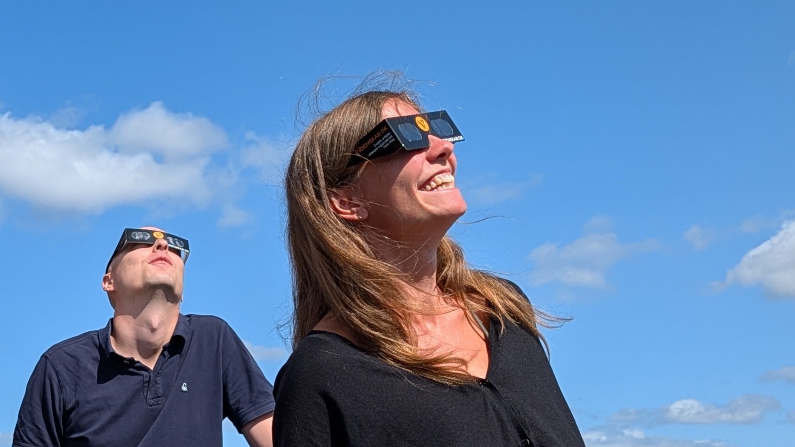 To personer, der bærer beskyttelsesbriller til solformørkelsen, kigger smilende op mod himlen under en klarblå himmel med spredte skyer – et ægte øjeblik af hverdagsvidenskab, hvor nysgerrighed møder forundring.