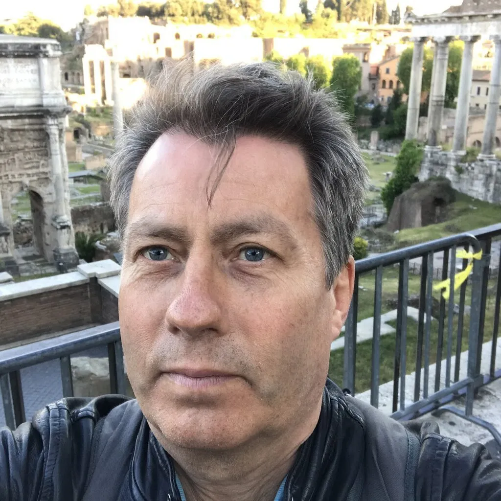En mand tager et selfie foran gamle romerske ruiner med stenbuer og søjler i det klare dagslys.