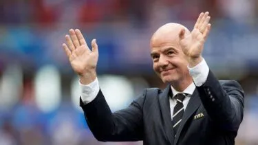 En mand i jakkesæt med et FIFA-badge løfter begge hænder og smiler, mens han står på et stadion med en sløret menneskemængde i baggrunden.