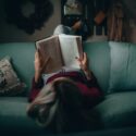 En person, der ligger på en blå sofa og holder en bog over ansigtet, mens vedkommende læser. Rummet er svagt oplyst, med en hyggelig indretning og en pude med håndskrift på.
