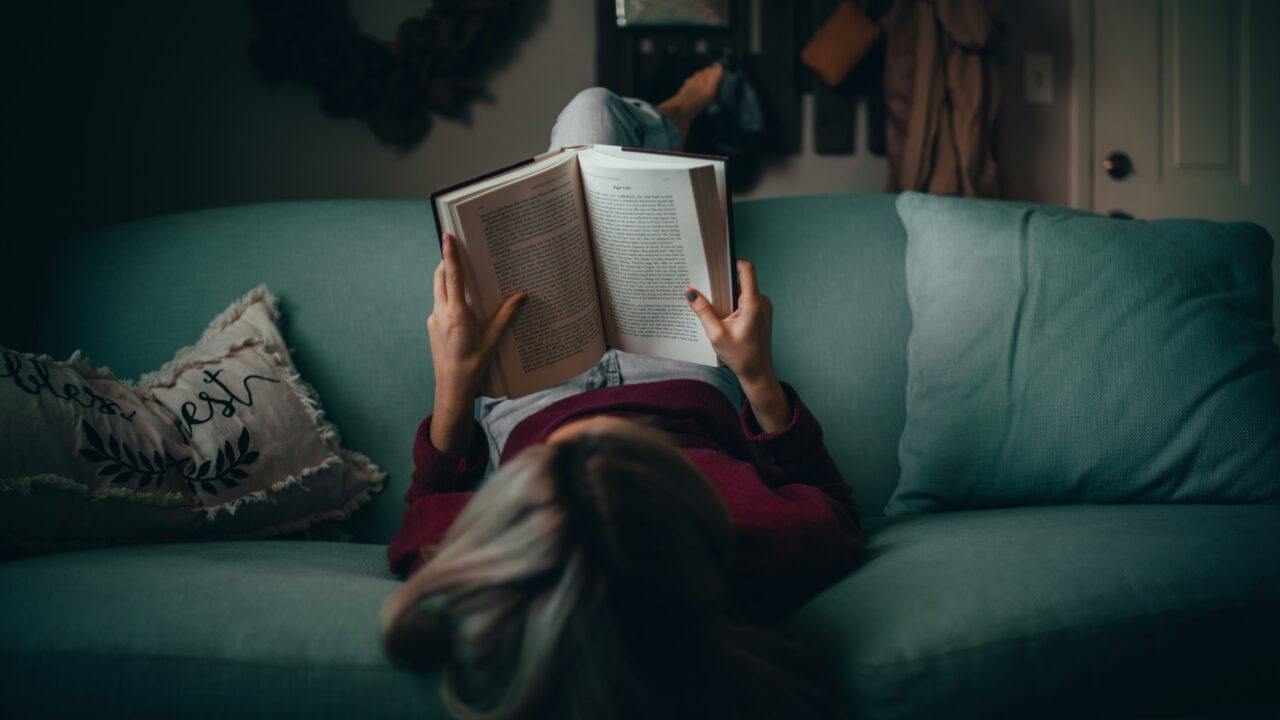 En person, der ligger på en blå sofa og holder en bog over ansigtet, mens vedkommende læser. Rummet er svagt oplyst, med en hyggelig indretning og en pude med håndskrift på.