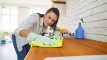 En person med handsker på rengør en køkkenbordplade af træ med en gul klud. I baggrunden ses en sprayflaske og et komfur.