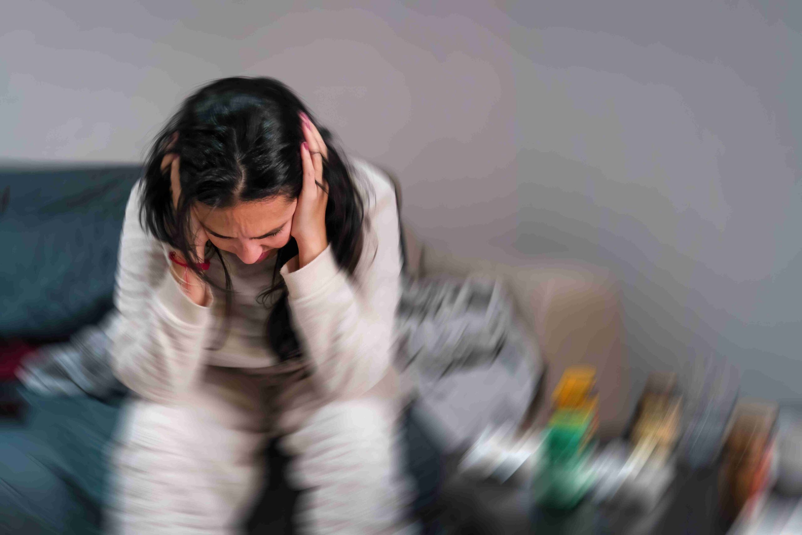 En kvinde sidder på en sofa og holder sig for hovedet i fortvivlelse. Billedet er sløret, hvilket understreger hendes følelsesmæssige uro. Der ligger medicin og papirlommetørklæder på et bord i nærheden.