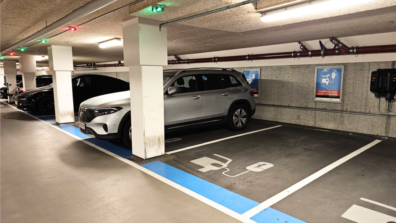 To elbiler holder parkeret ved ladestationerne i et indendørs parkeringshus. Gulvmarkeringerne og skiltene angiver de dertil indrettede ladepladser til elbiler. Området er godt oplyst af loftslamper.
