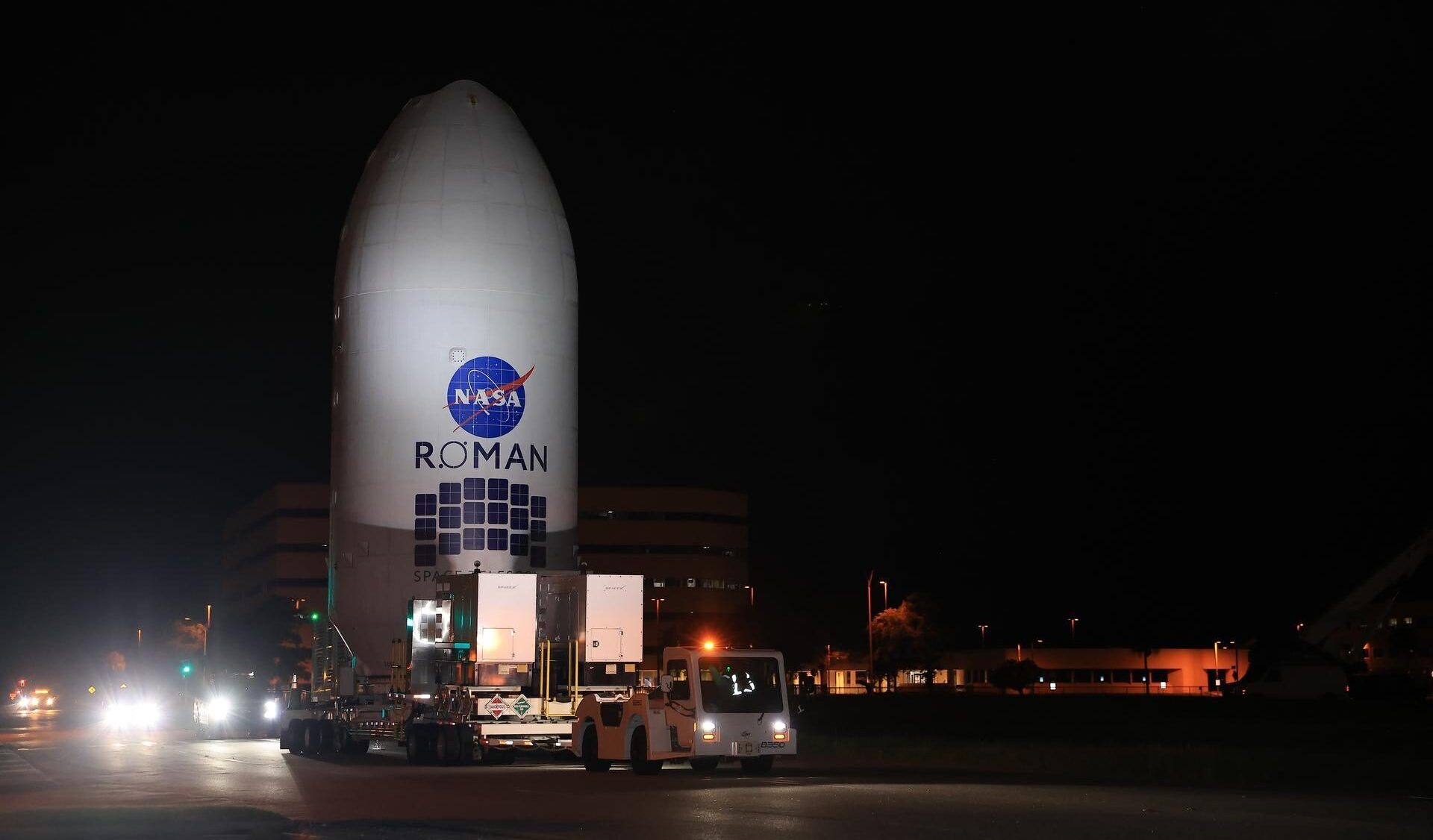 NASA's Roman-rumteleskop, der er dækket af en hvid beskyttelseskappe, transporteres om natten af en konvoj af køretøjer forbi oplyste bygninger.