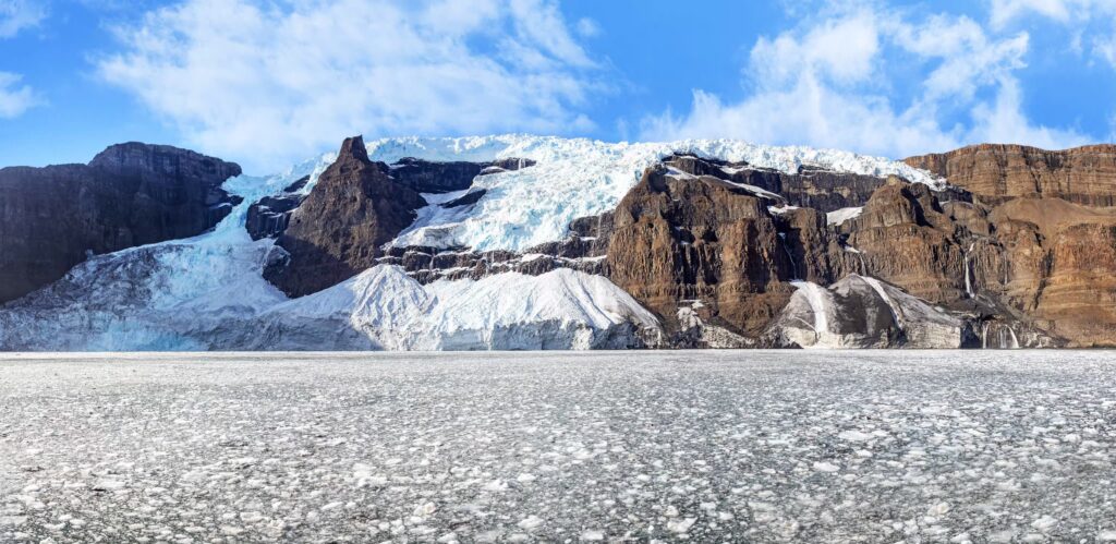 Et panorama af en gletscher med hvidt og blåt is på toppen af brune klipper under en strålende blå himmel, med en frossen, isdækket flade i forgrunden.