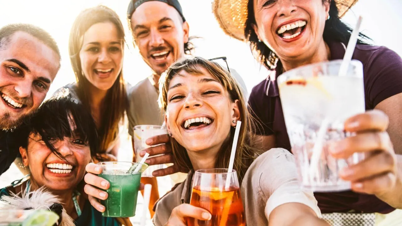 En gruppe venner, der smiler og holder farverige drinks i hånden, mens de tager et selfie udendørs i strålende solskin og ser glade og muntre ud.