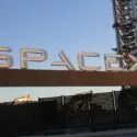 Et stort SpaceX-skilt i metal på en sort port, med entreprenørmaskiner og en høj bygning i baggrunden mod en klar blå himmel ved solnedgang.