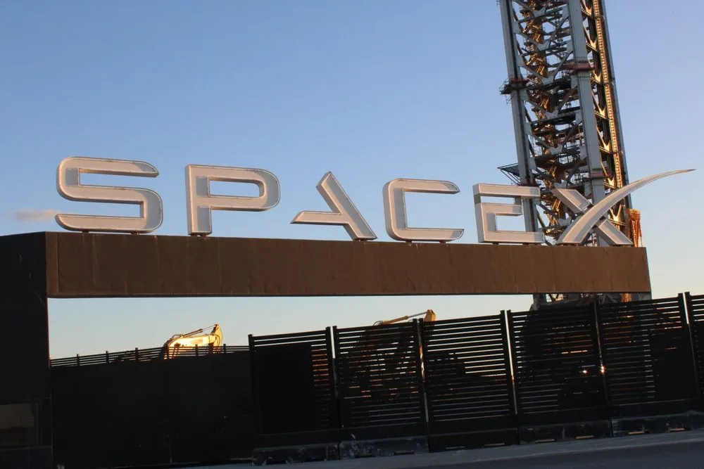 Et stort SpaceX-skilt i metal på en sort port, med entreprenørmaskiner og en høj bygning i baggrunden mod en klar blå himmel ved solnedgang.
