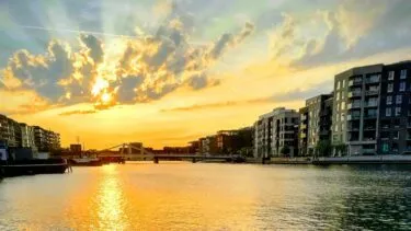 En farverig solnedgang over en rolig havnefront, med moderne lejlighedsblokke på begge sider og gyldent lys, der spejler sig i vandet. Skyer spredt ud over den klare himmel.