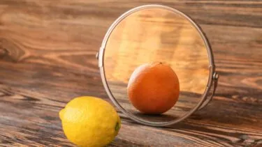 En citron ligger foran et rundt spejl på en træoverflade. Spejlet afspejler billedet af en appelsin i stedet for citronen.