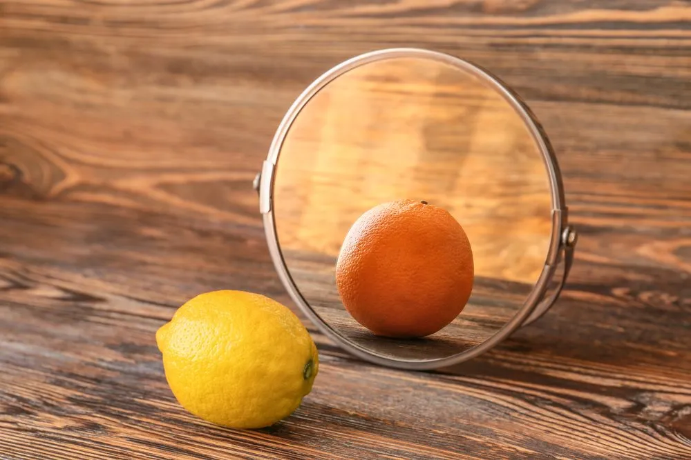 En citron ligger foran et rundt spejl på en træoverflade. Spejlet afspejler billedet af en appelsin i stedet for citronen.