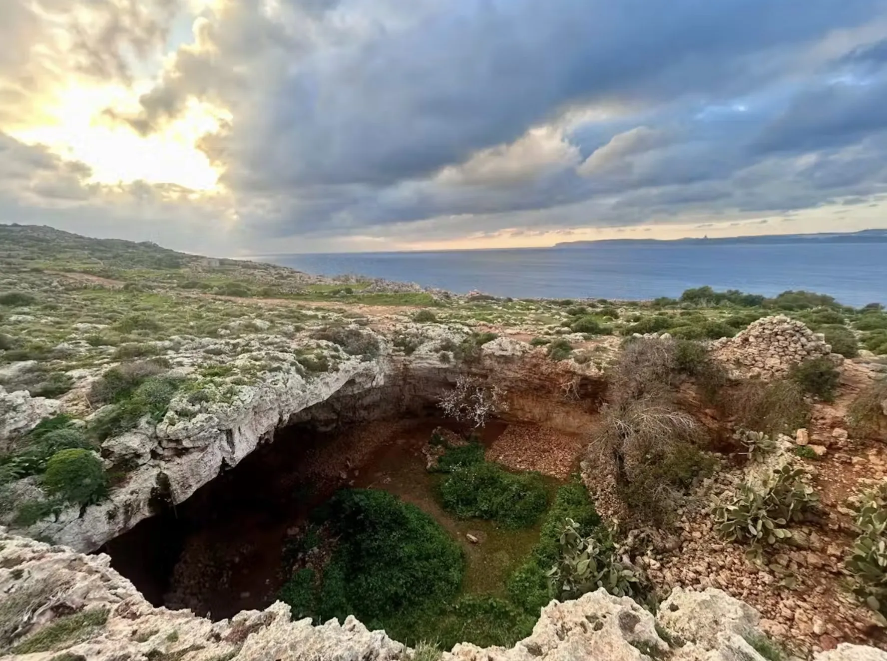 Et stort, klippefyldt hul, der er fyldt med grønne planter, ligger tæt på en forrevne kystlinje under en dramatisk, overskyet himmel ved solnedgang, med havet synligt i baggrunden.
