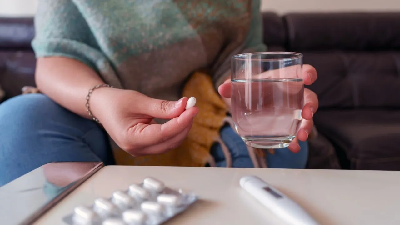 En person sidder på en sofa med en hvid pille i den ene hånd og et glas vand i den anden. På bordet foran vedkommende ligger en blisterpakning med piller og et termometer.