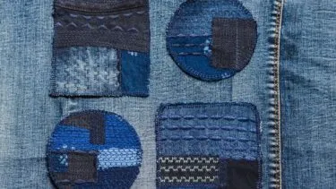 Fire blå stoflapper – to firkantede og to runde – er syet fast på denim. Hver lap har sin egen tekstur, syning og mønster i forskellige blå nuancer. Baggrunden er blåt denimstof med synlige sømme.