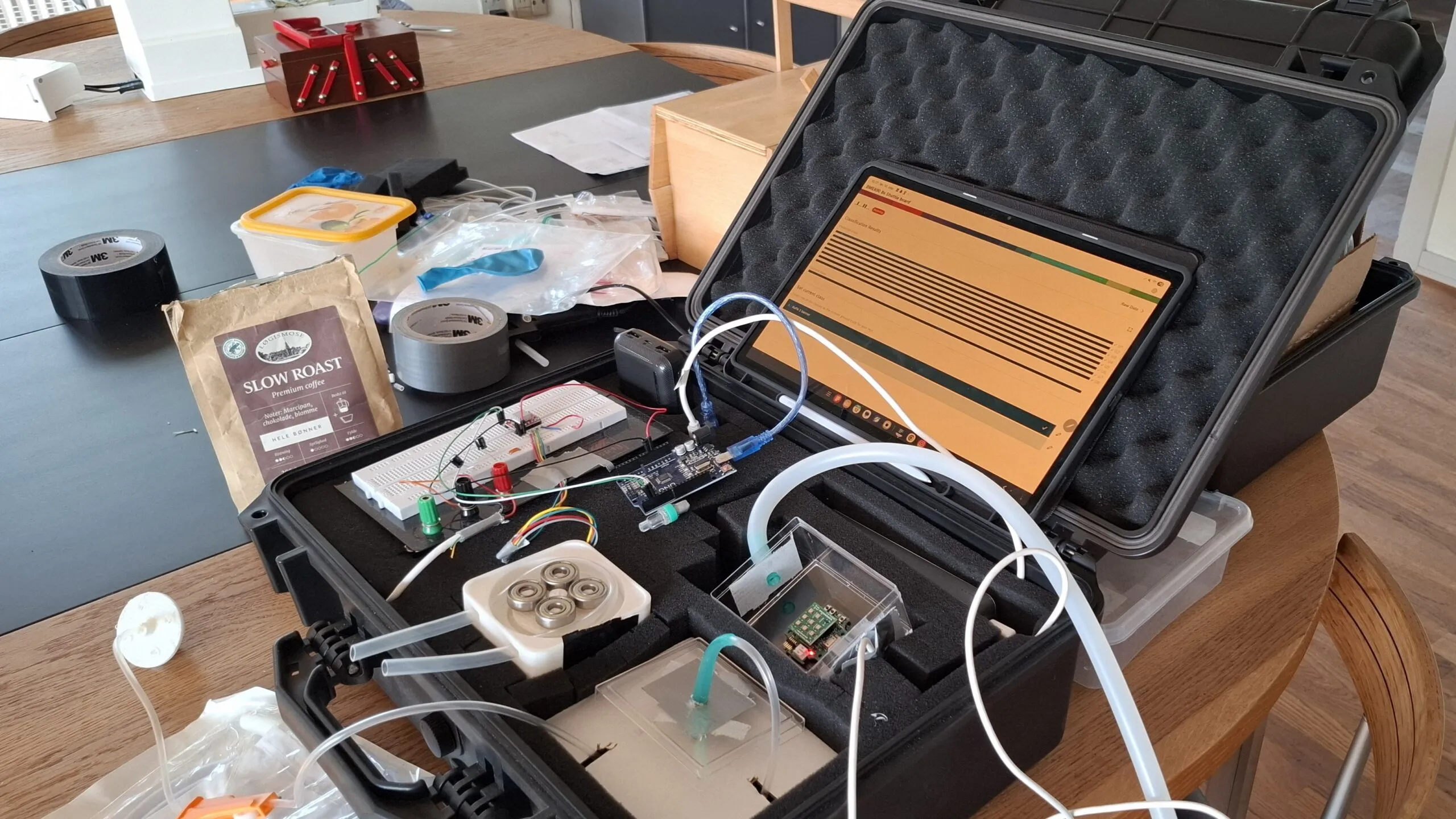 Et åbent, sort kabinet med elektroniske komponenter, rør, printkort og en tablet, der viser en graf, på et rodet bord med kaffeemballage, tape, ledninger og diverse små værktøjer spredt rundt omkring.
