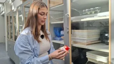 En kvinde i en kittel undersøger en lille beholder med rødt låg i et laboratorium, hvor hun står ved siden af hylder fyldt med bakker og laboratorieudstyr.
