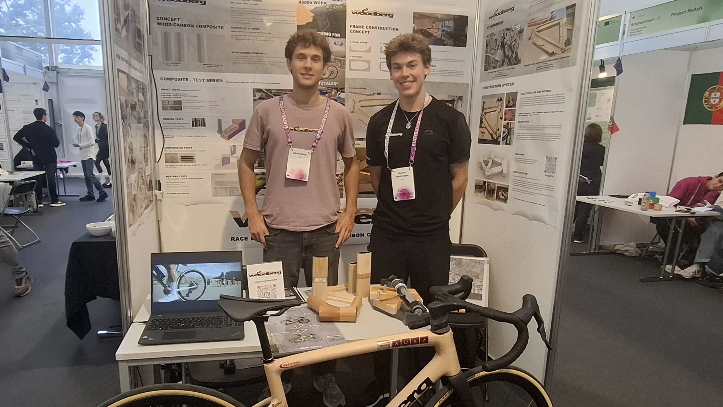 To unge mænd står og smiler bag en udstillingsstand, hvor der er en beige cykel, eksemplarer af cykelstel, en bærbar computer og informationsplakater – sandsynligvis på en videnskabs- eller innovationsmesse.