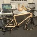 En lys træcykel er udstillet indendørs på et stativ foran et bord med en bærbar computer, træcykeldele og informationsmateriale. En person i grå bukser står i nærheden.