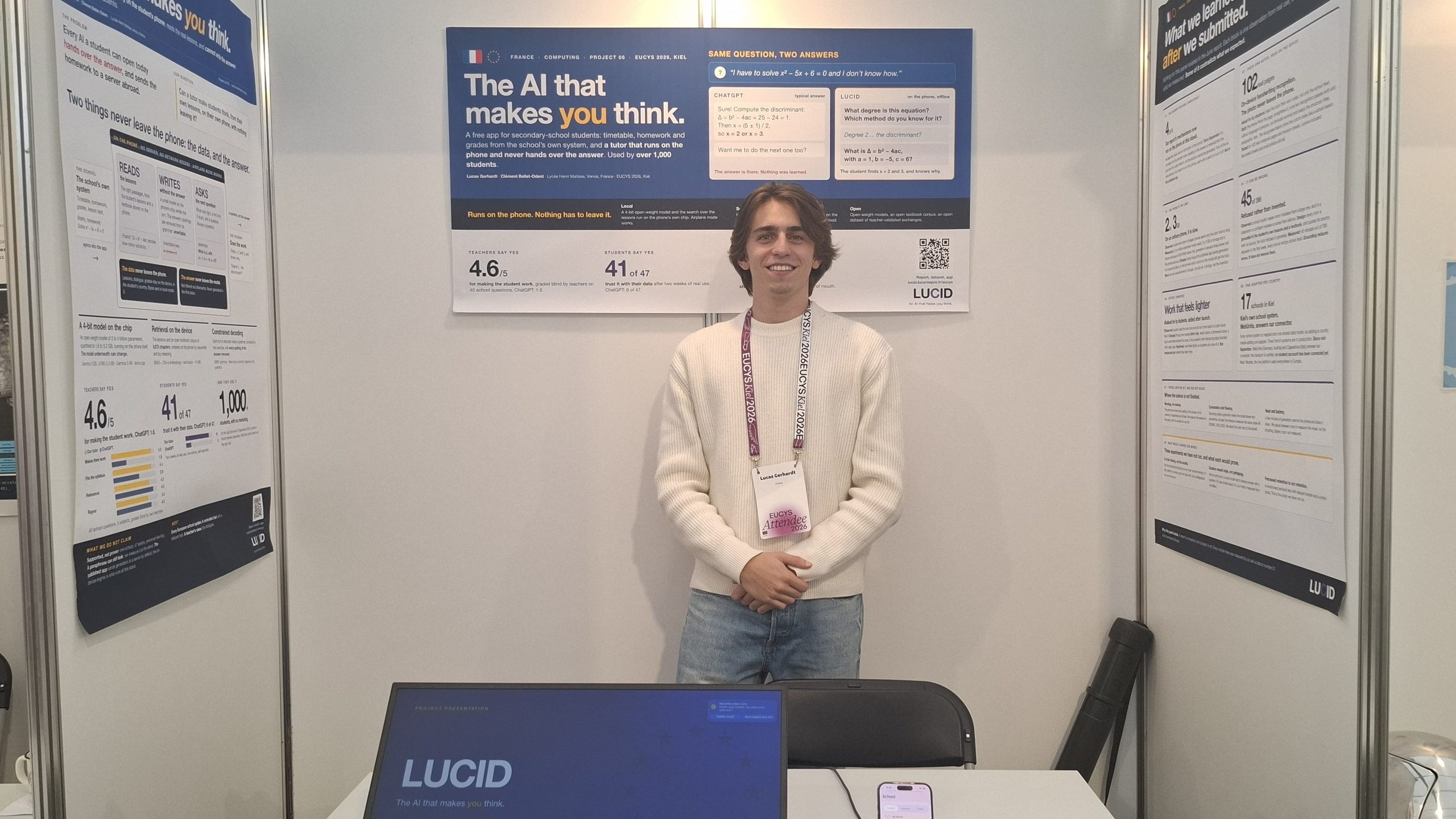 En person står og smiler ved en stand med plakater om AI-forskning. En bærbar computer med påskriften »LUCID« står på bordet foran vedkommende. Plakaterne indeholder tekst, diagrammer og oplysninger om kunstig intelligens.