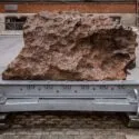 En stor, ru brun sten står udstillet på et robust metalstativ udendørs på en brostensbelagt overflade, med en murstensbygning og en gade i baggrunden.