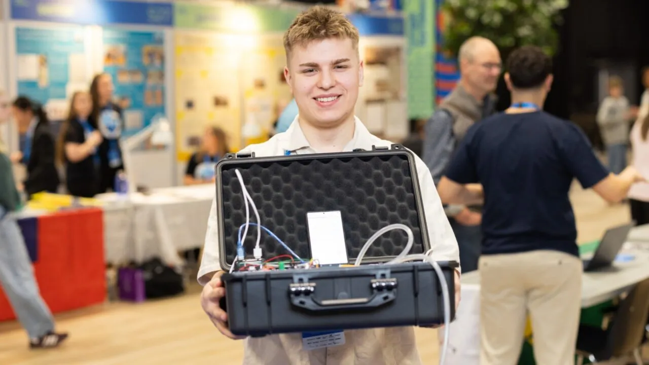 En ung person smiler, mens vedkommende holder en åben sort kuffert med elektronisk udstyr og kabler indeni, og står på et travlt indendørs arrangement med mennesker og informationsstande i baggrunden.