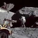 En astronaut i rumdragt står ved siden af en stor klippe på Månen, mens en månerover og udstyr kan ses i forgrunden under en mørk himmel.