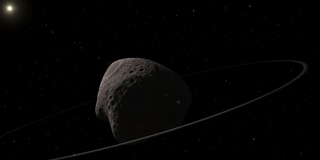 En grå asteroide med kraterer og et tyndt ringsystem kredser i rummet. Fjerne stjerner fylder den sorte baggrund, og solen lyser i det øverste venstre hjørne.