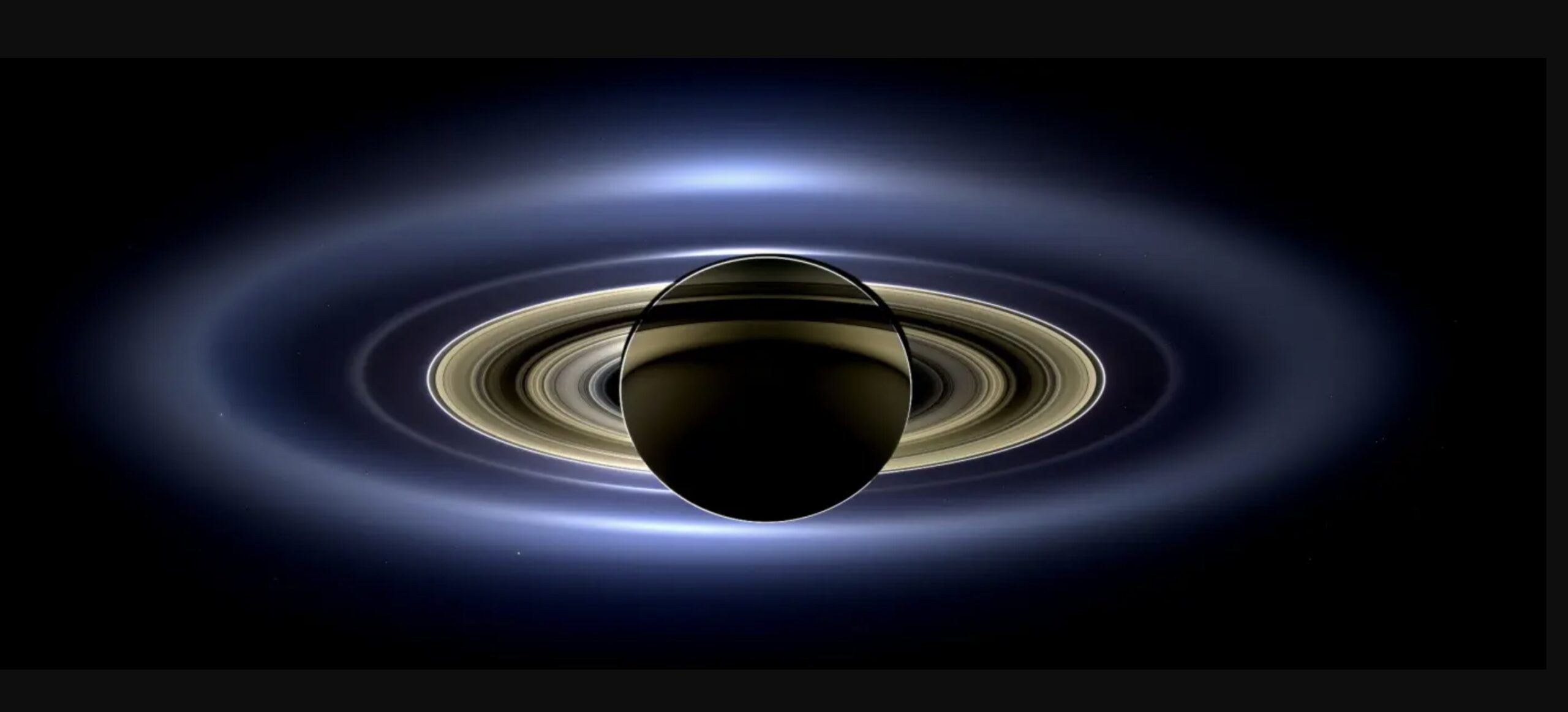 Et billede af Saturn set mod baggrunden viser planeten i skygge, mens dens ringe lyser op i koncentriske cirkler, omgivet af et svagt blåt skær mod rummets mørke.