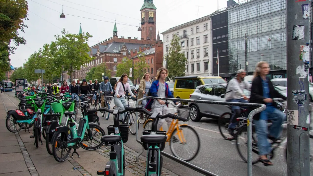 Folk cykler på en travl bygade, hvor der står parkerede cykler og scootere på begge sider, med biler og historiske bygninger i baggrunden. En smule bevægelsesslør antyder bevægelse.
