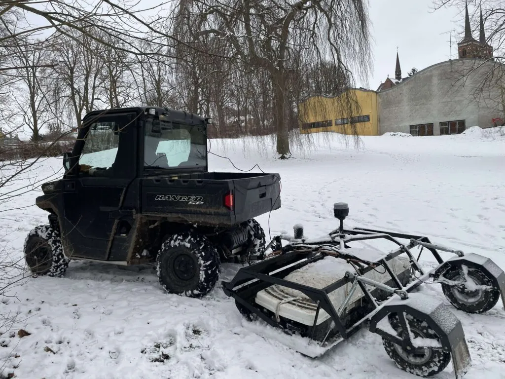 Et sort nyttekøretøj holder parkeret på en snedækket mark og trækker en anordning med metalramme. I baggrunden ses nøgne træer og snedækkede bygninger under en grå himmel.