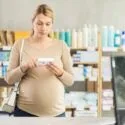 En gravid kvinde står i et apotek og holder en medicinæske, som hun læser på, mens man i baggrunden kan se hylder med produkter.