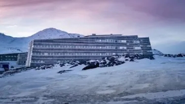 En moderne, fleretagers bygning, der er dækket af rim, står i et snedækket, klippefyldt landskab med bjerge i baggrunden under en farverig lyserød og lilla himmel.
