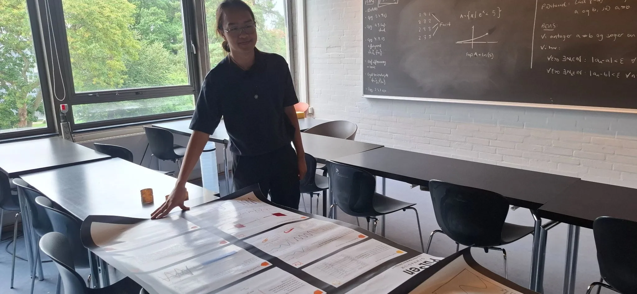En person står i et klasseværelse ved siden af et bord og viser en stor videnskabelig plakat med diagrammer og grafer; i baggrunden ses tavler med ligninger.