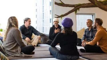 Seks personer sidder i en cirkel på puder i et lyst rum med store vinduer og et vægmaleri af en gren, hvor de deltager i en gruppediskussion eller meditation.