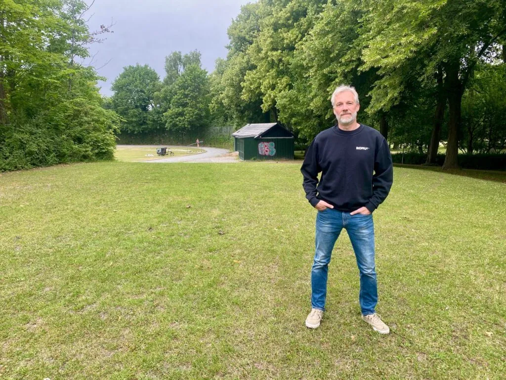 En mand står på en græsplæne med hænderne i lommerne. Bag ham ses træer, et lille skur med graffiti og en asfalteret sti under en overskyet himmel.