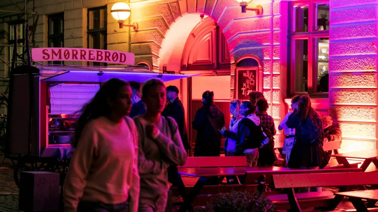 Om aftenen samles folk ved en smørrebrødsvogn uden for en bygning, der er oplyst af lyserøde og lilla lys; nogle går forbi i forgrunden, mens andre står og snakker ved vognen.