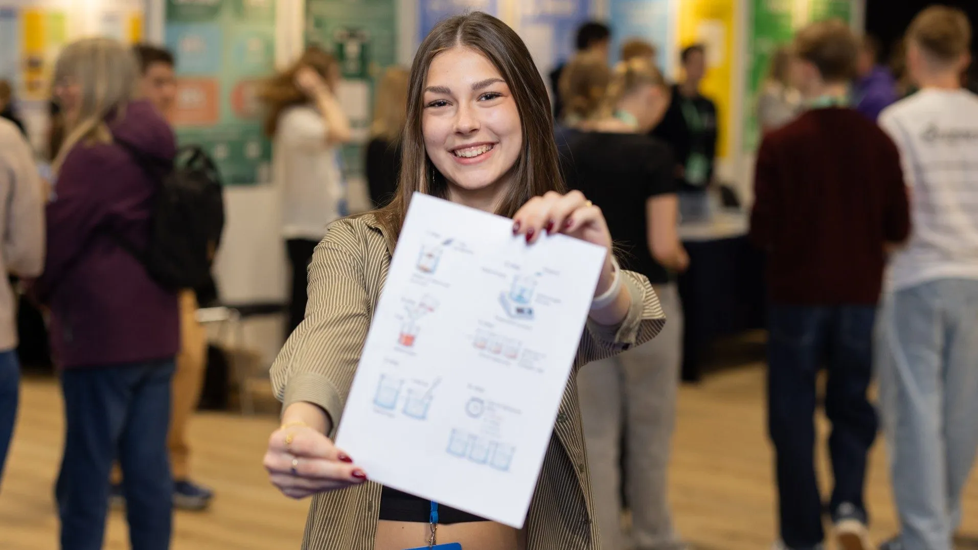 En smilende ung kvinde holder et udskrevet ark med diagrammer op mod kameraet ved et indendørs arrangement, hvor der er flere mennesker og farverige plakater i baggrunden.