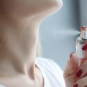 En person med røde negle sprøjter parfume på halsen og har en hvid bluse på. Baggrunden er let sløret.