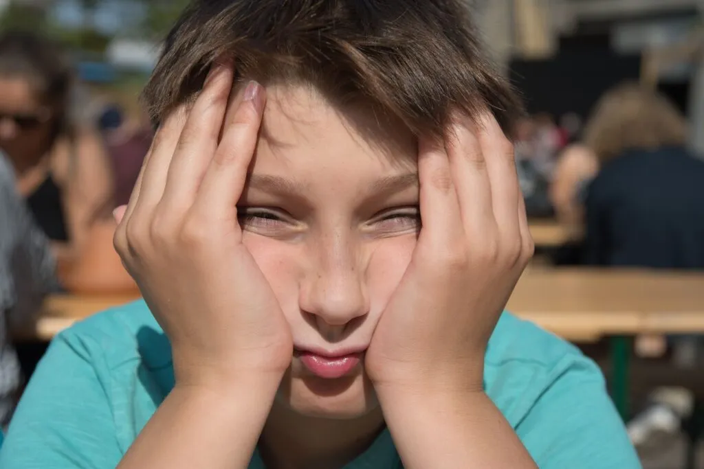 En dreng i en blågrøn skjorte sidder ved et bord udendørs, holder hænderne mod kinderne og ser direkte ind i kameraet med et udtryk, der tyder på kedsomhed eller træthed. Andre personer i baggrunden er slørede.