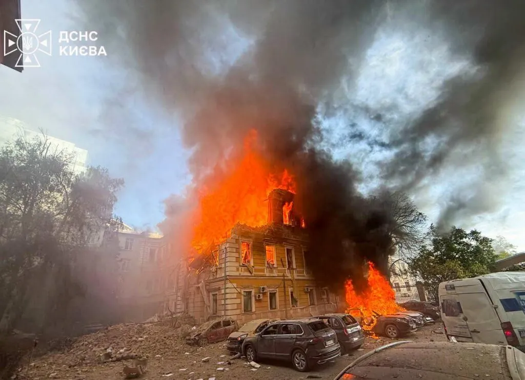 En stor bygning står i flammer og er omgivet af tæt, sort røg, mens der ligger vragdele spredt rundt omkring, og biler i nærheden er beskadiget. Der ses udrykningskøretøjer og officielle køretøjer. Himlen er delvist tilsløret af røg.