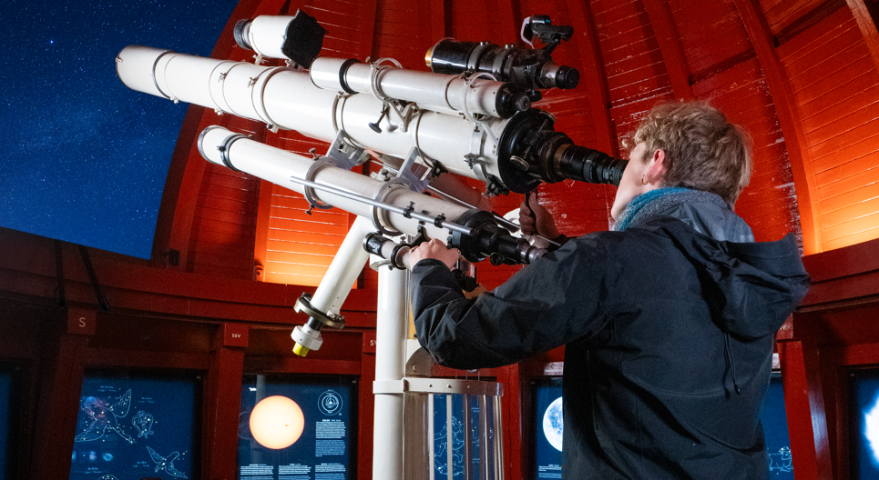 En person kigger gennem et stort teleskop inde i et observatorium med røde vægge og informationsskilte, mens nattehimlen kan ses gennem en åben kuppel.