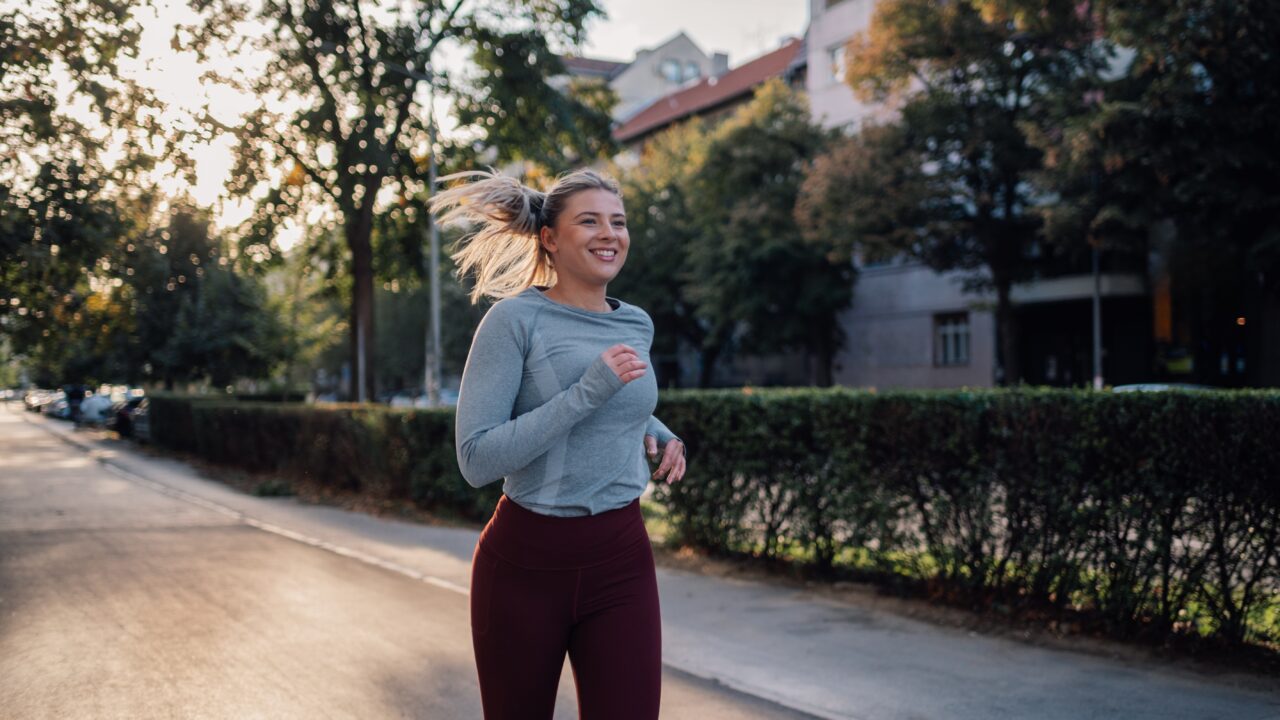 En kvinde i en grå langærmet bluse og rødbrune leggings løber på en træbeklædt bygade og smiler, mens solen går ned i baggrunden.