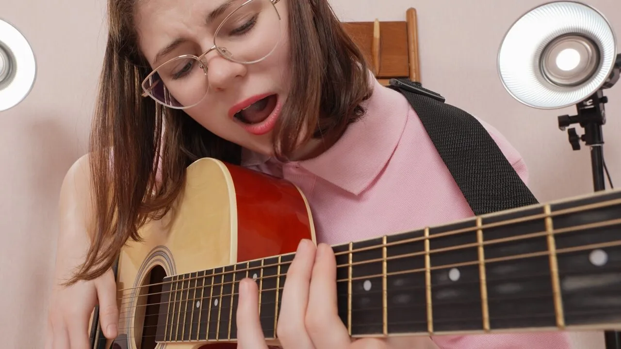 En ung kvinde med briller og en lyserød bluse spiller på en akustisk guitar og synger inderligt, mens der er sat studielys op bag hende.