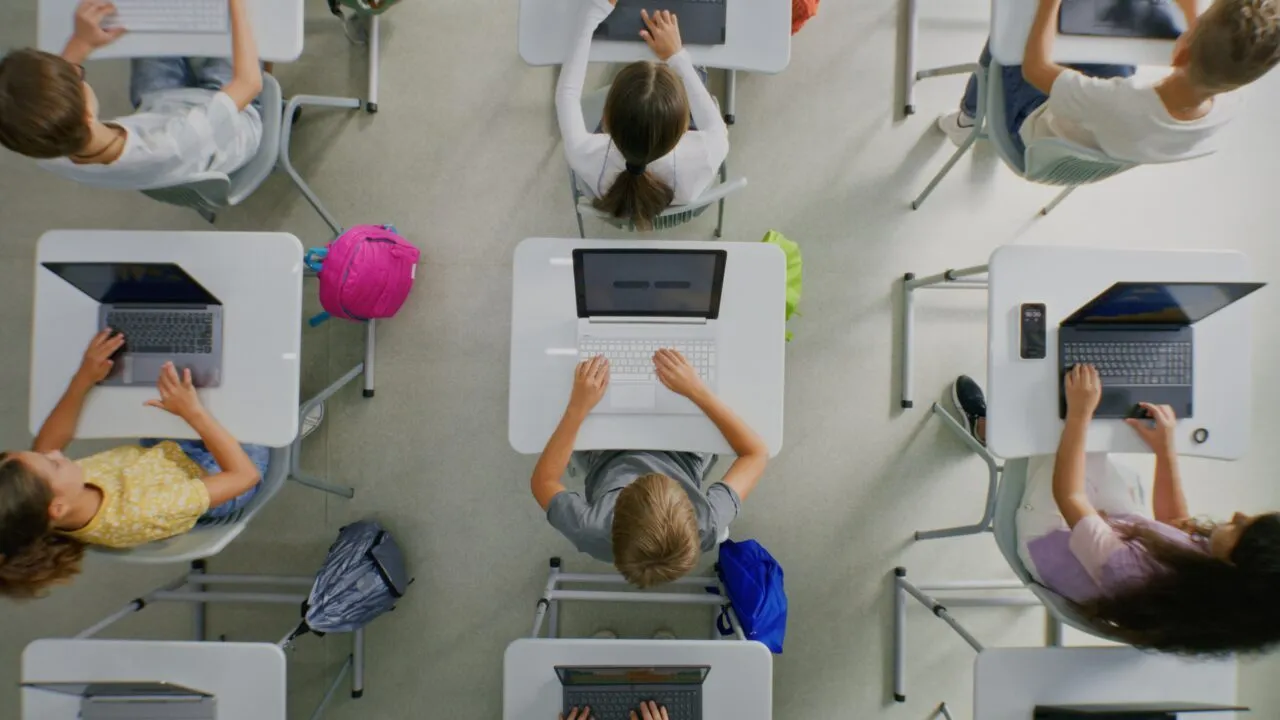 Set ovenfra: Elever, der sidder ved skriveborde og bruger bærbare computere i et klasseværelse. Rygsækkene står på gulvet ved siden af skrivebordene.
