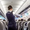 En stewardesse i uniform betjener passagererne inde i flykabinen, hvor man kan se rækker af siddende rejsende og skærme i loftet.