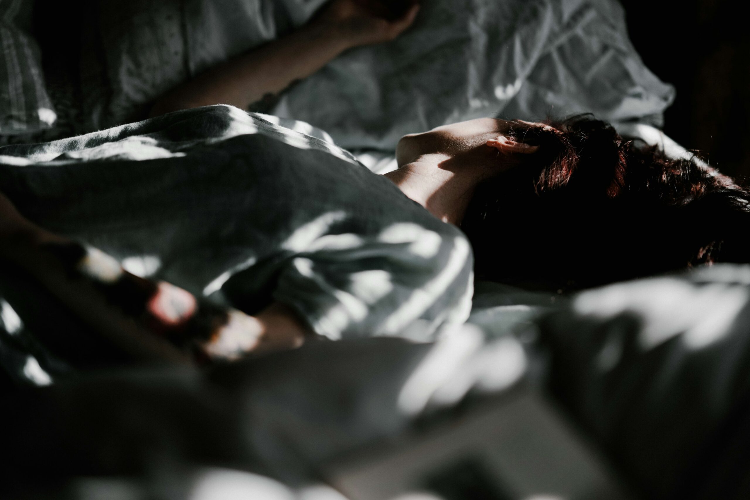En person, der ligger på en seng, delvist i skygge, iført en lys skjorte. Blødt sollys trænger ind og kaster mønstre på personens krop og sengetøjet. Stemningen er rolig og fredfyldt.
